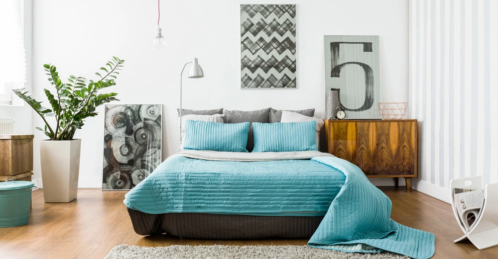 Bett mit blauem Überwurf steht in einem Schlafzimmer umrahmt von Holzmöbeln großen Kunstwerken einer Pflanze und dezenten Lampen auf einem hellen Holzboden mit weißen Wänden.