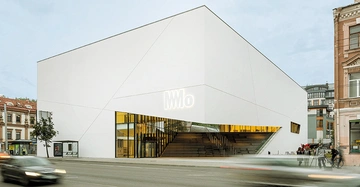 Ein großes weißes Gebäude mit der Beschriftung MMo an der Fassade Menschen gehen und fahren auf der Straße davor die Umgebung besteht aus anderen Gebäuden und einem Gehweg