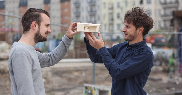 Zwei Männer halten ein Modellgebäude hoch und beobachten es genau auf einer Baustelle in einer urbanen Umgebung mit mehrstöckigen Wohngebäuden im Hintergrund.