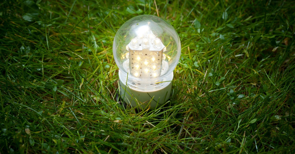 Eine leuchtende Glühbirne steht aufrecht im nassen Gras eines Gartens während der Dämmerung.