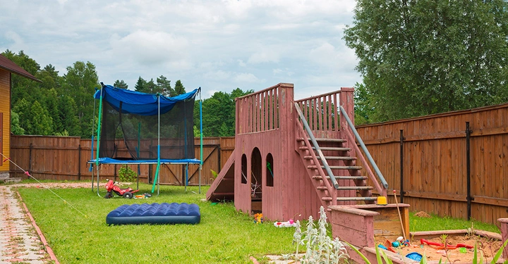 Ein rotes Holzspielhaus mit Treppe steht auf einem grünen Rasen in einem umzäunten Garten. Daneben befinden sich ein Trampolin, Spielzeuge und eine blaue Luftmatratze.