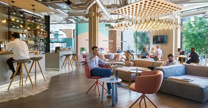 Galerie-Bild 2: Personen arbeiten und entspannen in einem modernen Café mit stilvollem Bartresen, gemütlichen Sofas und futuristischen Leuchten. Hohe Fenster bieten reichlich Tageslicht und einen Blick ins Grüne.