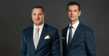 Zwei Männer stehen nebeneinander in eleganten Anzügen mit Krawatten in einem Studio vor dunkelgrauem Hintergrund und blicken direkt in die Kamera.