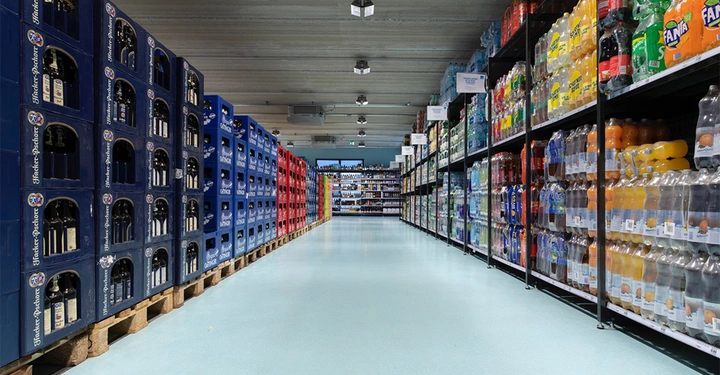 Galerie-Bild 5: Paletten mit Flaschen und Kisten sind gestapelt. Sie stehen in einem Getränkemarkt. Links sind Getränkekisten aus Holz und rechts sind Plastikflaschen auf Metallregalen.