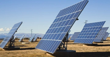 Solarmodule stehen winklig auf einer trockenen Wiese und sind in Reihen angeordnet. Im Hintergrund sind Strommasten und Berge zu sehen unter einem klaren blauen Himmel.