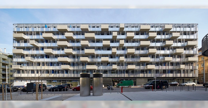 Galerie-Bild 15: Ein modernes Gebäude mit mehreren Balkonen; davor parken Autos und Lieferwagen am Straßenrand; im Vordergrund befinden sich zwei große zylindrische Lüftungsschächte auf einem Platz.