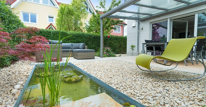 Ein grüner Liegestuhl steht auf einem Kiesweg neben einem kleinen Teich mit Schilf im Garten eines Hauses mit gelber Fassade und großen Fenstern.