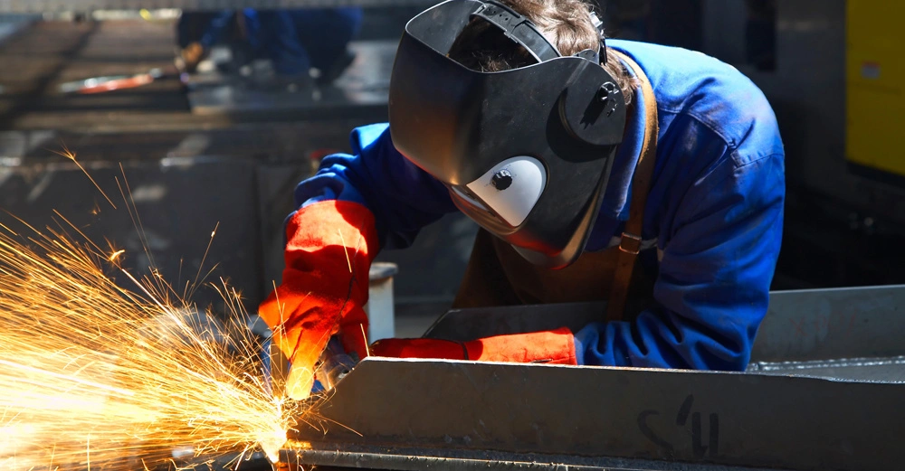 Ein Arbeiter trägt eine Schweißmaske und Handschuhe und schleift Metall mit einem Winkelschleifer in einer Werkstatt. Funken fliegen in verschiedene Richtungen. Anderer Arbeiter im Hintergrund.