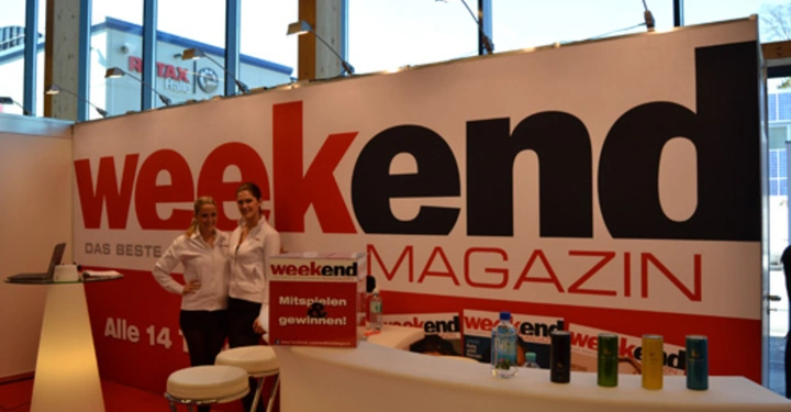 Galerie-Bild 30: Zwei Personen stehen an einem Messestand Weekend Magazin Mitspielen und Gewinnen im Hintergrund Weitsee durch die Fenster beleuchteter Raum mit Stühlen und einem Laptop auf einem hohen Tisch