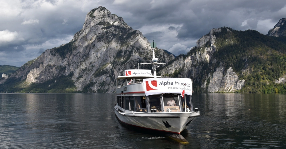 Ein kleines Passagierschiff fährt auf einem See mit umliegenden hohen Bergen. Auf dem Schiff steht alpha innotec the better way to heat. Der Himmel ist bewölkt und dramatisch.