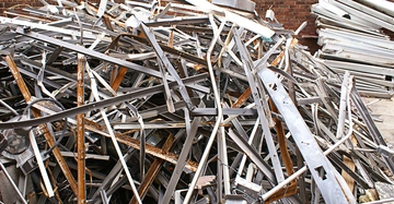 Metallteile liegen auf einem großen Haufen. Einige Teile sind verrostet und andere scheinen neu zu sein. Im Hintergrund sind Ziegelwände und weitere Metallhaufen zu sehen, die den Hof einrahmen.