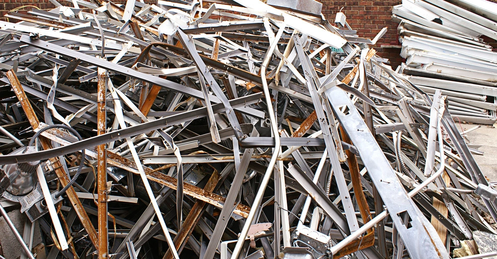 Metallteile liegen auf einem großen Haufen. Einige Teile sind verrostet und andere scheinen neu zu sein. Im Hintergrund sind Ziegelwände und weitere Metallhaufen zu sehen, die den Hof einrahmen.