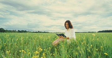 Eine Frau sitzt und liest ein Buch in einem weiten grünen Feld mit gelben Blumen unter einem bewölkten Himmel.