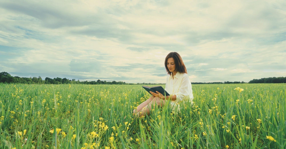 Eine Frau sitzt und liest ein Buch in einem weiten grünen Feld mit gelben Blumen unter einem bewölkten Himmel.