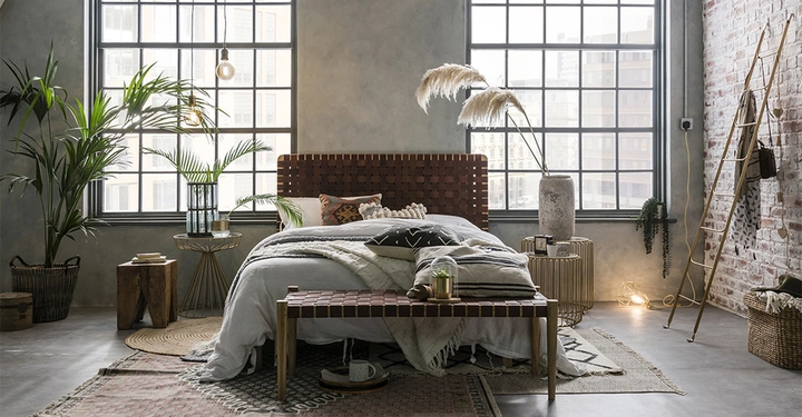 Ein Bett mit Kissen und Decken steht in einem industriell eingerichteten Schlafzimmer mit großen Fenstern hohen Pflanzen rustikalen Möbeln und gemischten Textilien. Ein hölzerner Hocker und eine Leiter sind daneben.