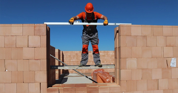 Galerie-Bild 2: Bauarbeiter balanciert Wasserwaage auf roten Ziegelmauern Baustelle Klarer blauer Himmel Ziegelsteine und Baumaterialien im Hintergrund