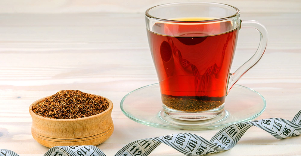 Ein Glas Tee steht auf einem Untersetzer mit einer Holzschale voller getrockneter Teeblätter daneben Ein Maßband schlängelt sich um die beiden Objekte Der Hintergrund ist ein heller Holztisch