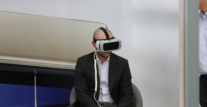 Galerie-Bild 9: Ein Mann sitzt auf einem Stuhl und trägt ein Virtual-Reality-Headset in einem modernen Büroraum, während eine andere Person teilweise sichtbar im Hintergrund steht.