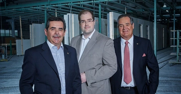 Drei Männer stehen nebeneinander und lächeln in die Kamera. Sie tragen formelle Kleidung. Der Hintergrund zeigt eine industrielle Umgebung mit Metallstrukturen und Arbeitsbereichen.