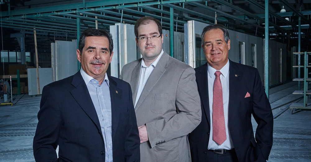 Drei Männer stehen nebeneinander und lächeln in die Kamera. Sie tragen formelle Kleidung. Der Hintergrund zeigt eine industrielle Umgebung mit Metallstrukturen und Arbeitsbereichen.