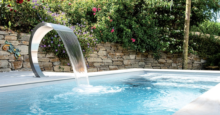Wasser fließt aus einem modernen Wasserfalldurchlauf in einen Pool in einem gepflegten Garten mit einer Steinmauer im Hintergrund und blühenden Pflanzen darüber.