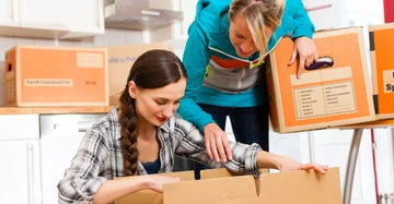 Zwei Frauen packen Kartons in einer Küche aus Umzugskartons mit Beschriftungen wie Küche Wohnzimmer und 40 kg umgeben sie. Eine Frau kniet über einen offenen Karton die andere hält einen geschlossenen.