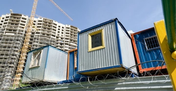 Containerboxen stehen aufeinander gestapelt hinter einem Stacheldrahtzaun, während daneben ein unfertiges, hohes Gebäude und ein gelber Baukran sichtbar sind. Der Himmel ist klar und blau.