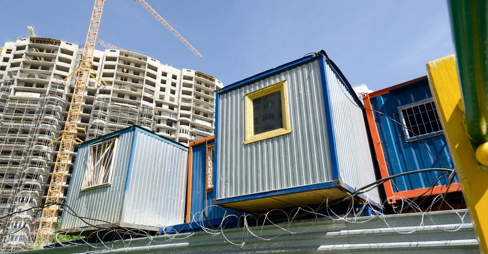 Containerboxen stehen aufeinander gestapelt hinter einem Stacheldrahtzaun, während daneben ein unfertiges, hohes Gebäude und ein gelber Baukran sichtbar sind. Der Himmel ist klar und blau.