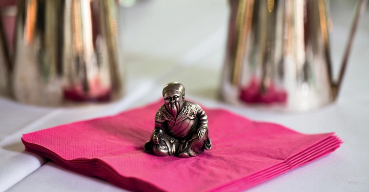 Galerie-Bild 3: Eine kleine Metallfigur sitzt auf einer pinken Serviette; im Hintergrund sind zwei silberne Kaffeekannen auf einem Tisch zu sehen.