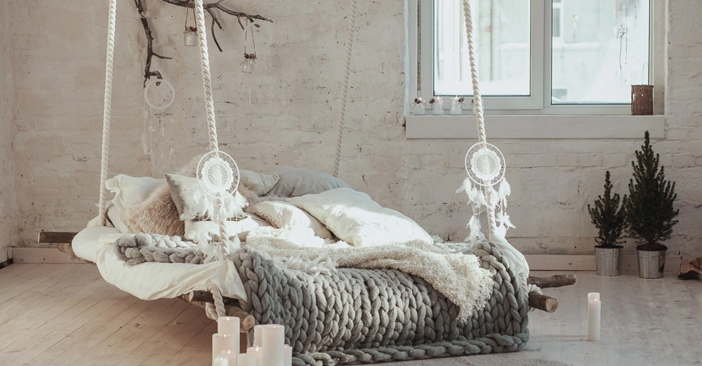 Ein hängendes Bett aus Seilen und Holzstämmen ist mit Decken und Kissen dekoriert in einem Zimmer mit weißen Backsteinwänden Kerzen Pflanzen und einem großen Fenster dekoriert.