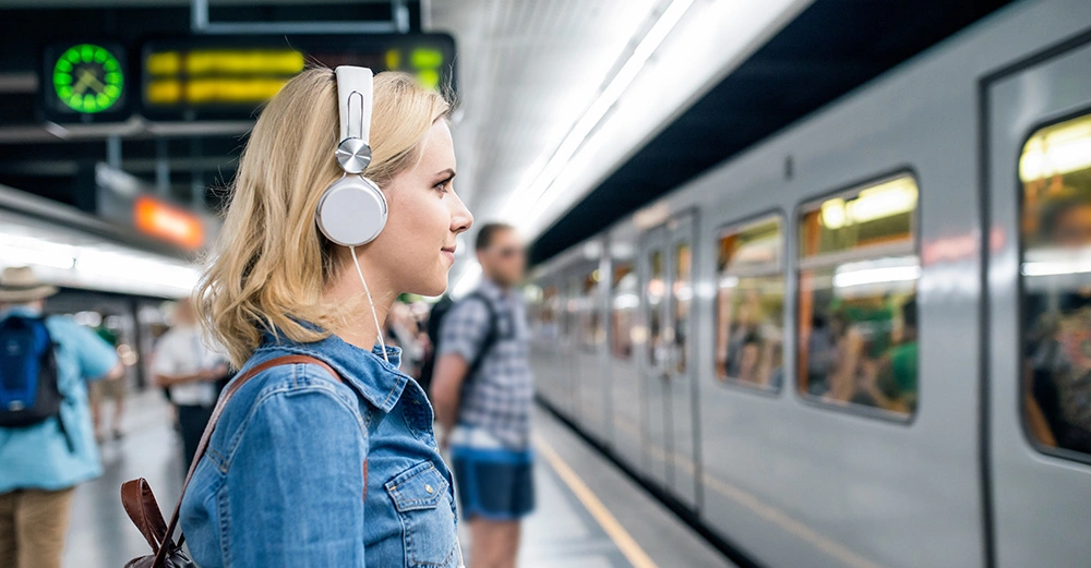 Frau trägt Kopfhörer und schaut auf einen vorbeifahrenden Zug in einer U-Bahn-Station mit anderen Reisenden und einer Uhr im Hintergrund.
