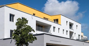 Modernes Mehrfamilienhaus mit weißer Fassade und gelben Akzenten im obersten Stockwerk, mehreren Balkonen und großen Fenstern unter blauem Himmel; im Vordergrund steht ein grüner Baum.