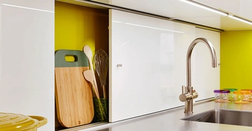 Ein Küchenarmatur steht über einem Edelstahlspülbecken. Links davon sind ein halb offener Schrank mit Schneidebrettern und Kochutensilien sowie bunte Gläser auf der Arbeitsplatte. Hintergrund gelb.