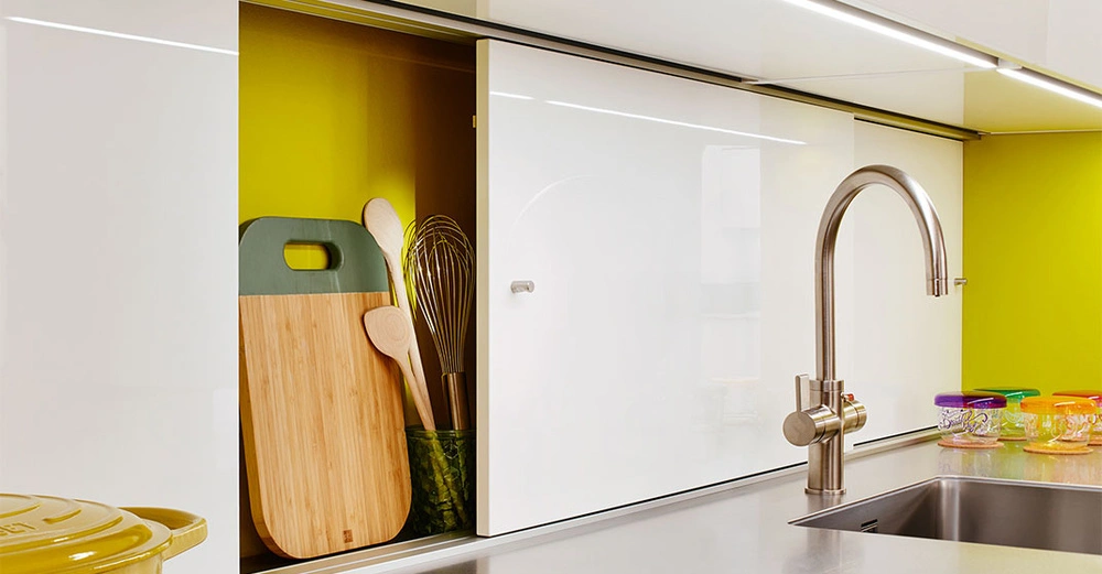 Ein Küchenarmatur steht über einem Edelstahlspülbecken. Links davon sind ein halb offener Schrank mit Schneidebrettern und Kochutensilien sowie bunte Gläser auf der Arbeitsplatte. Hintergrund gelb.