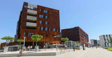 Mehrstöckiges modernes Gebäude mit rostfarbenen Fassaden Menschen laufen und sitzen in einem sonnigen gepflasterten Platz umgeben von anderen modernen Gebäuden und kleinen Bäumen unter einem klaren blauen Himmel