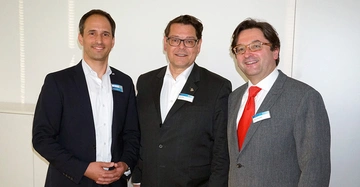 Drei Männer stehen nebeneinander lächeln in die Kamera und tragen Namensschilder in einem hell erleuchteten Raum mit weißen Wänden. Die Namensschilder sind leider nicht lesbar.
