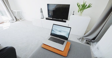 Ein Laptop liegt auf einem Tisch vor einem Fernseher in einem modernen, minimalistisch eingerichteten Wohnzimmer mit cremefarbenem Teppich, weißen Vorhängen und Pflanzen auf einem weißen Schrank.