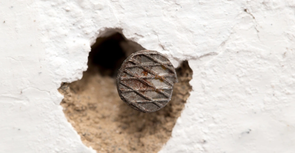 Nagel steht aus bröckelnder weißer Wand heraus, umgeben von beschädigtem Putz und freigelegtem Mauerwerk.