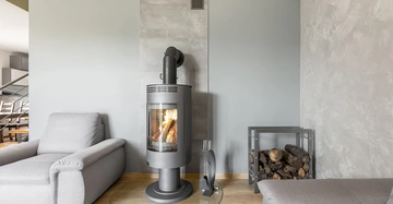 Ein moderner Kaminofen brennt auf einem runden Glasboden in einem Wohnzimmer mit grauen Wänden umgeben von zwei grauen Sesseln und einem Holzregal.