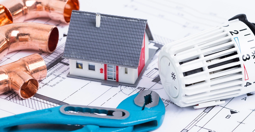 Ein Tisch mit Modellhaus, Kupferrohren einem Heizregler und einer blauen Zange, die über Bauplänen liegen.