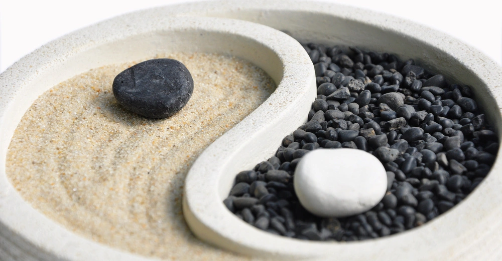 Ein Yin-Yang-Symbol aus Sand und schwarzen Steinen mit einem schwarzen Stein auf der Sandseite und einem weißen Stein auf der Steineseite in einem runden flachen Behälter.