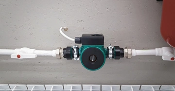 grüne Umwälzpumpe frontal mit weißen Heizungsrohren, die rechts und links von der Pumpe wegführen. Dahinter eine Sichtbetonwand