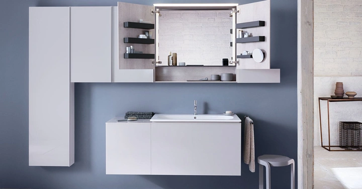 Spiegelschrank mit offenen Türen zeigt Regale mit Toilettenartikeln. Waschbecken mit Handtuch daneben steht davor. Im Hintergrund befinden sich dunkelgraue Wände und ein angrenzender Raum.