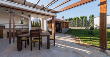 Ein Holztisch mit vier Stühlen steht auf einer gefliesten Terrasse unter einem teilweisen Holzpergola. Im Hintergrund befindet sich eine gepflegte Rasenfläche und ein überdachter Grillbereich.