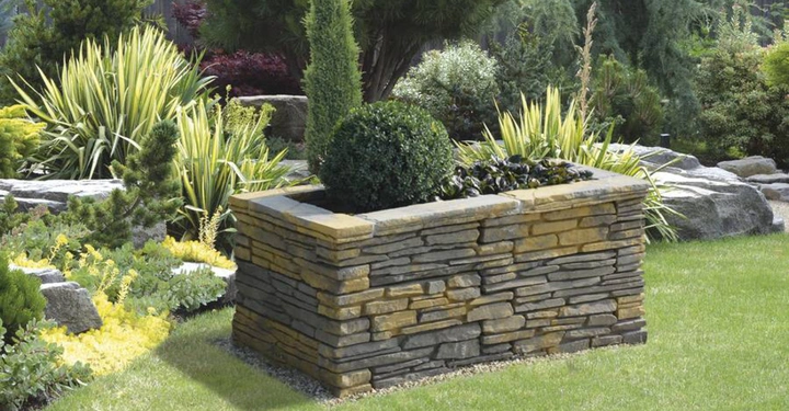 Galerie-Bild 12: Rechteckiger Steinpflanzer hält kugelförmigen Strauch und hohen schmalen Baum umgeben von eckigen Steinen und dichten grünen Pflanzen in einem gepflegten Garten