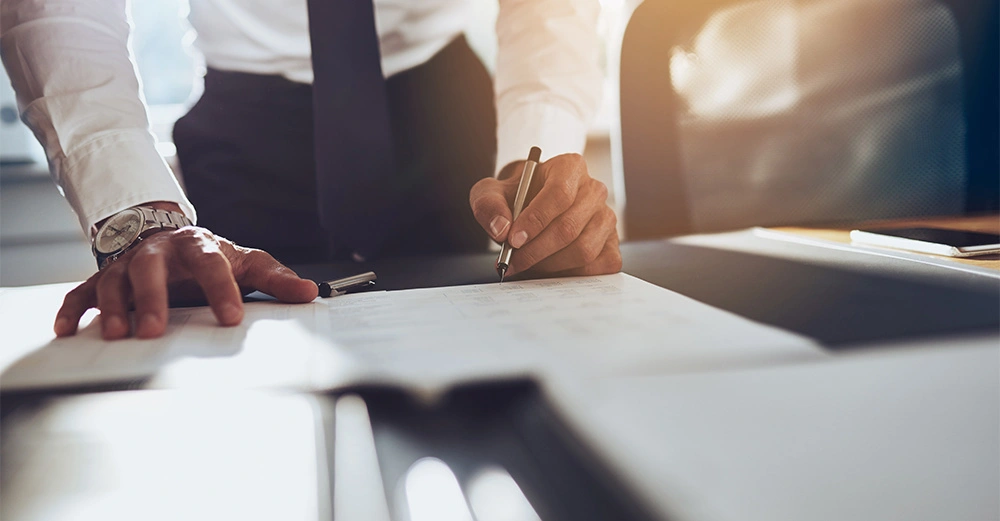 Person schreibt etwas auf Papier, steht dabei über einem Schreibtisch in einem Büro mit Sonnenlicht im Hintergrund. Ein in der Nähe liegender Stuhl und weitere Büromaterialien sind erkennbar.