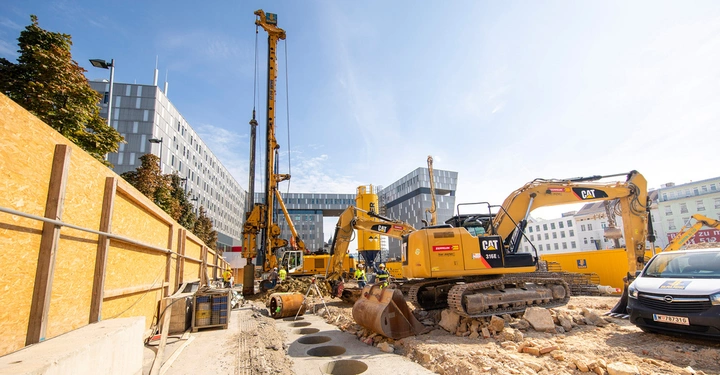 Galerie-Bild 4: Bagger arbeiten auf einer Baustelle umgeben von hohen Gebäuden und einem Bauzaun bei klarem Himmel. Schilder am Bagger: CAT Zeppelin. Nummernschild auf dem Van: W 787731 G