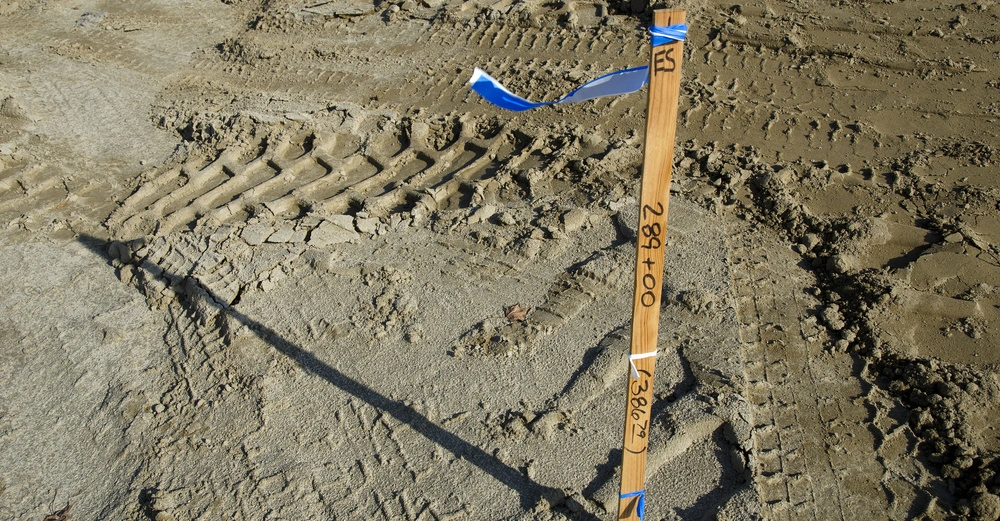 Holzpflock steht im Boden mit blauen Bändern befestigt und hat die Aufschrift "ES 289+00 3862.7" im Kontext einer Baustelle mit Erde und Reifenspuren.