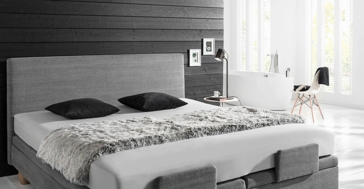 Graues Bett mit zwei schwarzen Kissen steht vor schwarzer Holzvertäfelung auf der linken Seite Sessel, Lampe und Badewanne in hellem Raum mit großen Fensterläden im Hintergrund