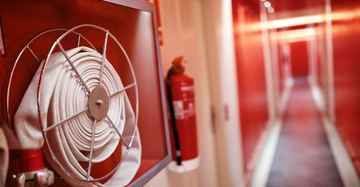 Ein aufgerollter Feuerwehrschlauch und ein Feuerlöscher befinden sich in einem roten Korridor. Links befindet sich ein Feuerlöscher an einer weißen Wand. Der Hintergrund zeigt einen unscharfen, langen Flur.